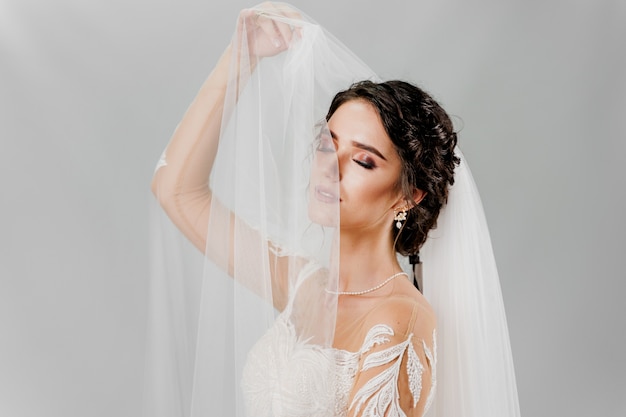 Young attractive bride in wedding dress touches bridal veil on white wall in studio. 