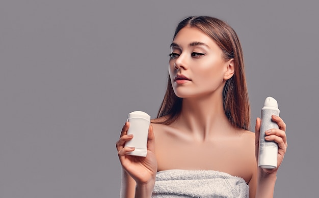 Young attractive brunette girl with loose hair with deodorant stick and antiperspirant aerosol isolated on gray background. Body care.