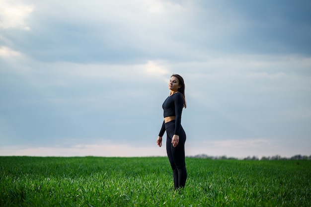 Young attractive brunette woman standing in nature, wearing black leggings and a black top. Summer day, green grass. Athletic body and healthy lifestyle.