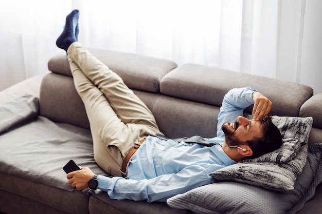 Young attractive businessman lying on couch at home, relaxing and using smart phone.