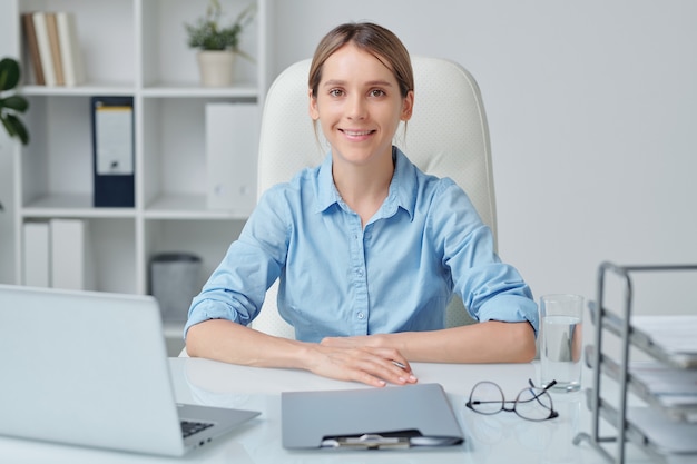 Young attractive businesswoman with blond hair and toothy smile looking at you while sitting by desk and working in office