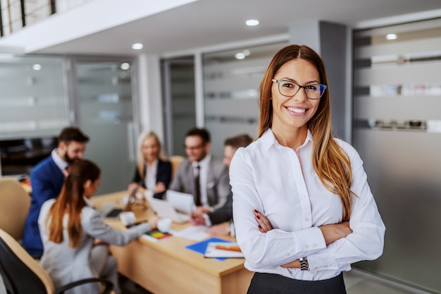 Young attractive Caucasian businesswoman in formal wear standing in boardroom with hands crossed and looking at camera. In background are her colleagues working on project.