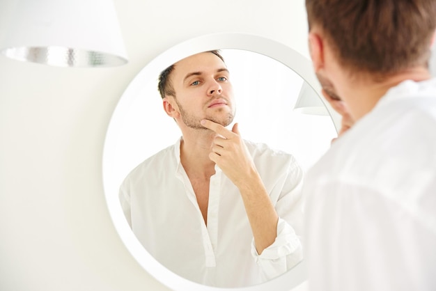 Young attractive Caucasian man examines his face in the mirror closeup Skin care concept