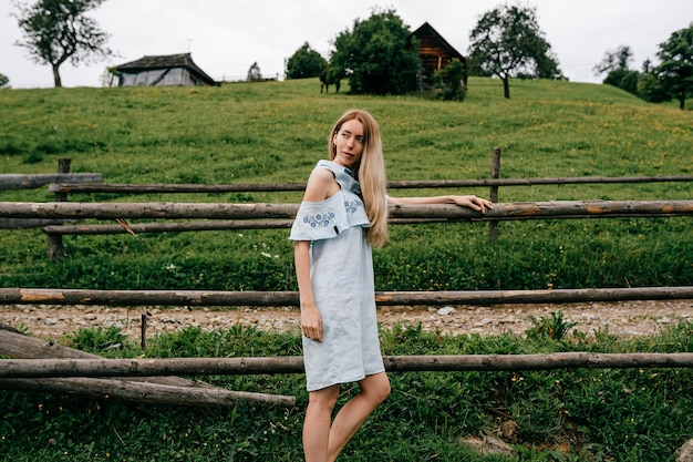 Young attractive elegant blonde girl in blue romantic dress posing near fence in the countryside