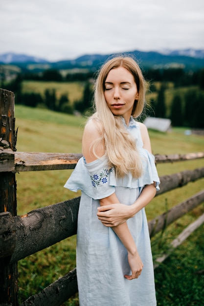 Young attractive elegant blonde girl in blue romantic dress posing near fence in the countryside