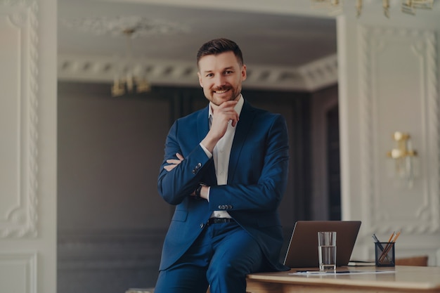 Young attractive executive manager or businessman in formal dark blue suit smiling and being confident while sitting on work desk with laptop, pencils and glass of water in stylish luxury office