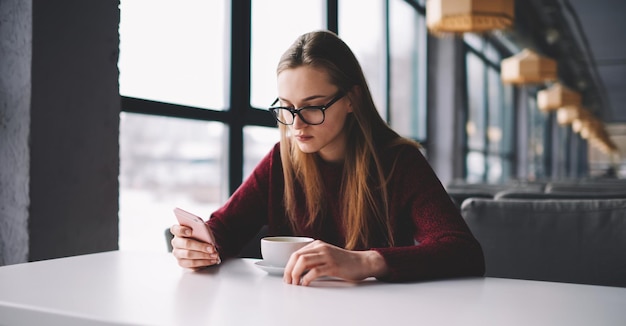 Young attractive female student sitting at favourite cafe and chatting with friends via smartphone