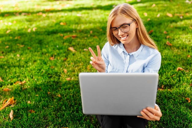 Young attractive girl in glasses, sitting with a laptop, talking on Skype and showing two fingers, on a laptop screen, in a park on a green lawn