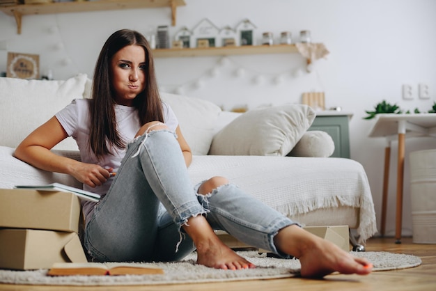 young attractive girl pouted her lips looks at camera and sits in front of sofa among boxes parcels