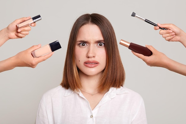 Young attractive girl with dark hair doing makeup being surrounded with hands with brushes for makeup expressing shock and negative emotions