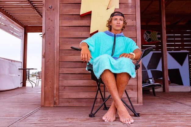 Young attractive guy surfer wearing a bathrobe is sitting on the terrace by the sea relaxing and looking out into the distance at sea