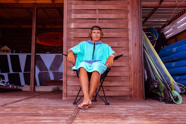 Young attractive guy surfer wearing a bathrobe is sitting on the terrace by the sea relaxing and looking out into the distance at sea