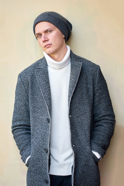 Young attractive hipster trendy man wearing a gray coat, white sweater and black jeans standing near wall