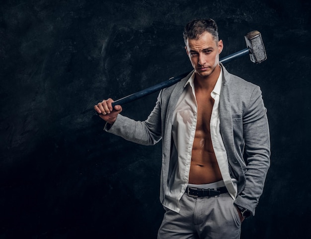 Young attractive hot man in suit and opened white shirt is holding long hammer in hand.