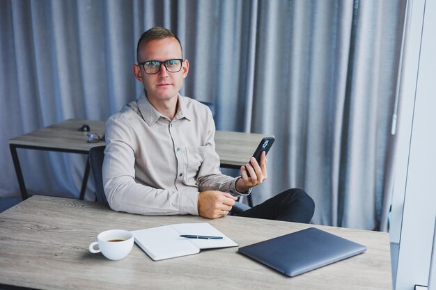 A young attractive male freelancer in glasses sits in the office and looks at the phone Handsome blogger writing ideas on phone while sitting at wooden table in office with laptop