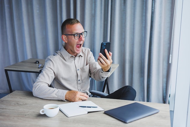 A young attractive male freelancer in glasses sits in the office and looks at the phone Handsome blogger writing ideas on phone while sitting at wooden table in office with laptop