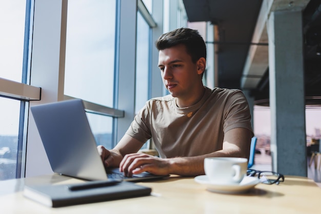 A young attractive male freelancer sits in the interior of a coffee shop and looks out the window Handsome blogger writing ideas in laptop while sitting at wooden table in cafe with laptop