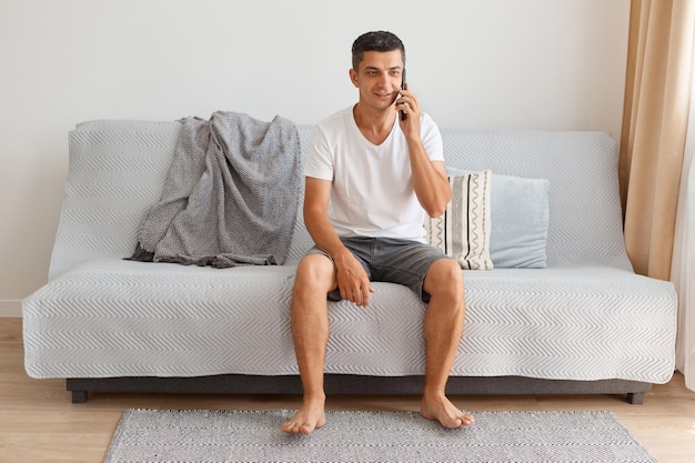 Young and attractive man talking on smart phone at home while sitting on cough in living room, wearing white t shirt and jeans short, looking away with positive facial expression.