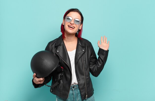 Young attractive red hair woman smiling happily and cheerfully, waving hand, welcoming and greeting you, or saying goodbye. motorbike rider concept