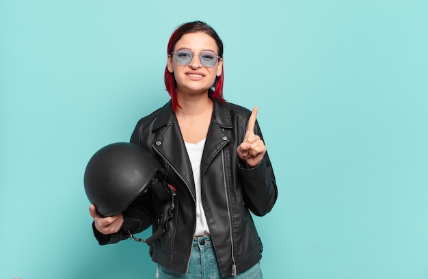 Young attractive red hair woman smiling and looking friendly, showing number one or first with hand forward, counting down. motorbike rider concept