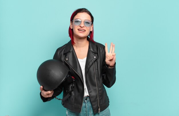 Young attractive red hair woman smiling and looking friendly, showing number three or third with hand forward, counting down. motorbike rider concept