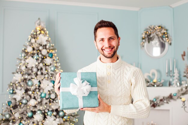 Young attractive smiling man posing in studio beautifully decorated for Christmas holding blue square gift box with white ribbon Winter holidays concept