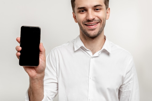 Young attractive smiling man in white shirt showing cellphone Focus on mobile close up isolated