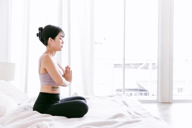 Young attractive sporty Asian woman practicing yoga on the bed doing Ardha Padmasana exercise meditating in Half Lotus pose with namaste indoor working out at home wearing sportswear