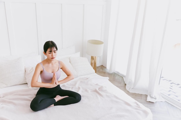 Young attractive sporty Asian woman practicing yoga on the bed doing Ardha Padmasana exercise meditating in Half Lotus pose with namaste indoor working out at home wearing sportswear