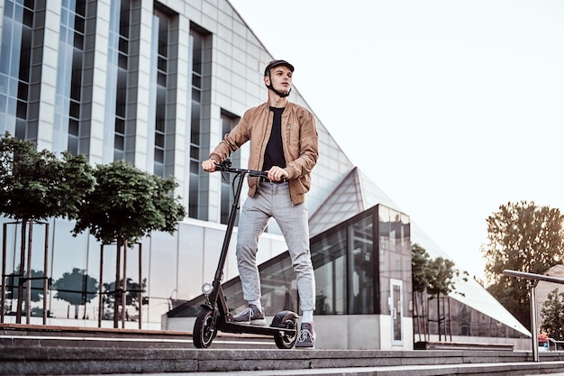 Young attractive student is driving his scooter neat interesting glass building at bright sunny day.