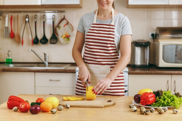 The young attractive woman in an apron cuts vegetables for salad with a knife in the kitchen. Dieting concept. Healthy lifestyle. Cooking at home. Prepare food.