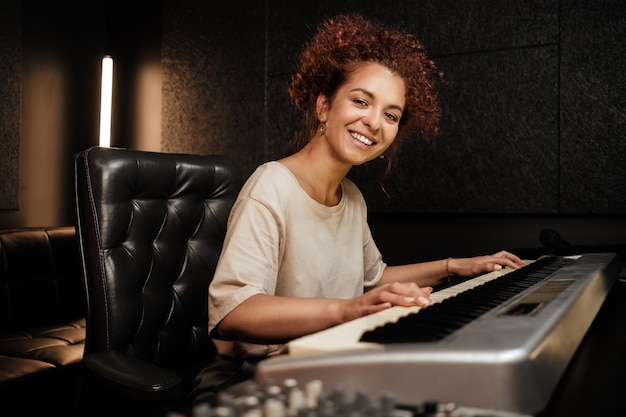 Young attractive woman happily looking in camera playing on electric piano in sound recording studio. Female musician working in studio