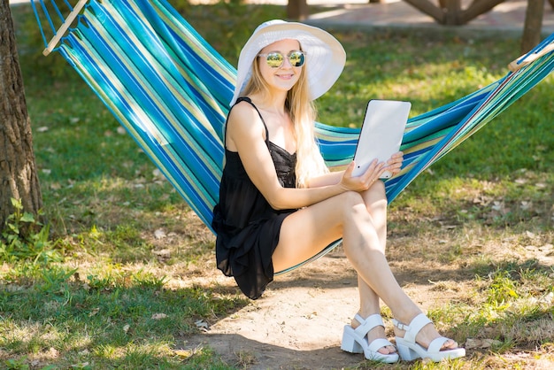 Young attractive woman in hat using a tablet on the hammock in the garden