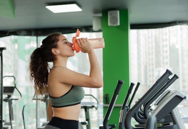 Young attractive woman is exercising on an elliptical exercise machine and drinking water from a bottle in the gym. Fitness, healthy lifestyle concept.