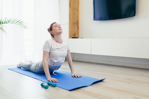 Young attractive woman is practicing yoga online using a mobile phone or smartphone app. Female yogi morning in casual clothes at home in living room, workout lotus position meditates on a mat