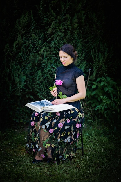 Young attractive woman sitting in a garden with a big book and a rose flower in her hands