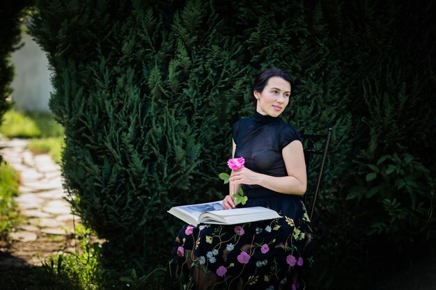 Young attractive woman sitting in a garden with a big book and a rose flower in her hands