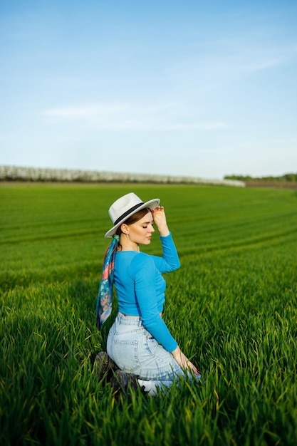 Young attractive woman in white hat blue shirt blue jeans posing in summer green field Copy empty space for text