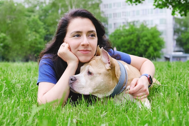 Young attractive woman with dark long hair lies on the lawn smiles and hugs the dog