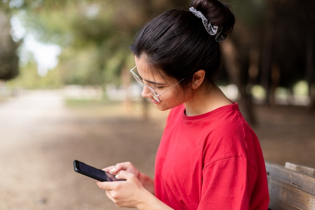 Young attractive woman with glasses sitting in a bench and using her phone in a park, with a red shirt. Seville, Spain