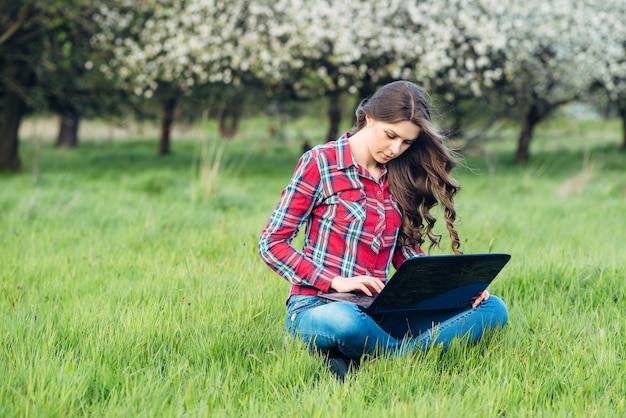 Young attractive woman with laptop on the grass in blooming garden