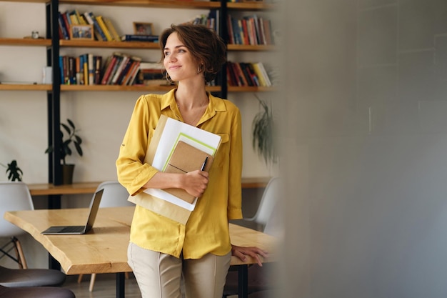 Young attractive woman in yellow shirt leaning on desk with notepad and papers in hand while dreamily looking aside in modern office