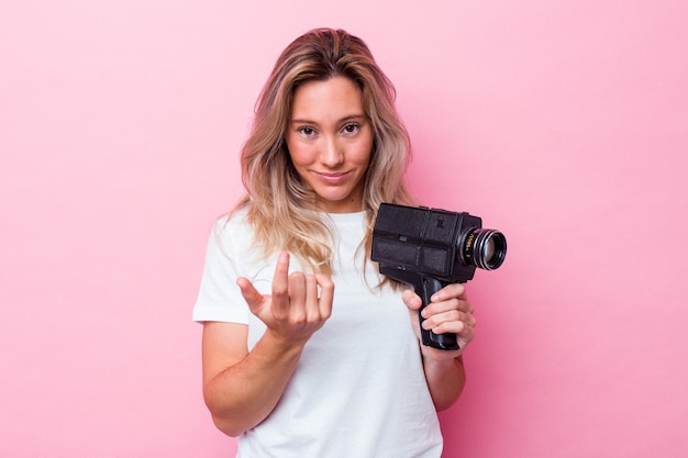 Young australian woman filming with a vintage video camera isolated pointing with finger at you as if inviting come closer.