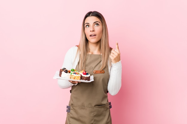 Young baker woman holding sweets pointing upside with opened mouth.