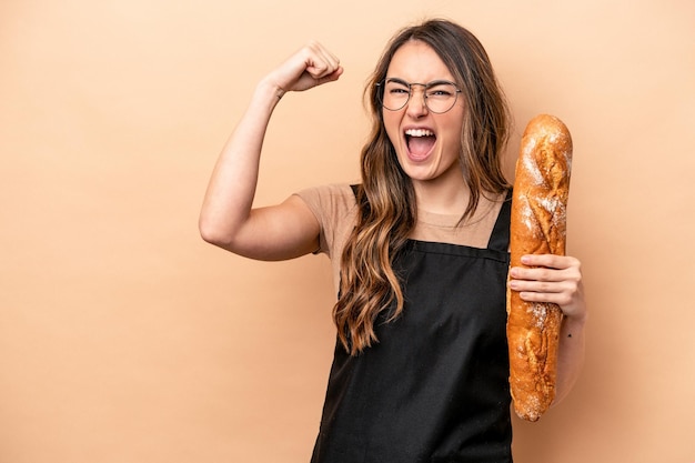 Young baker woman isolated on beige background raising fist after a victory winner concept