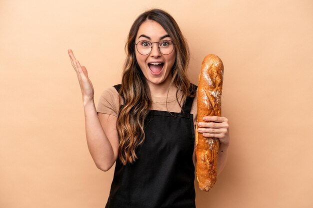 Young baker woman isolated on beige background receiving a pleasant surprise excited and raising hands