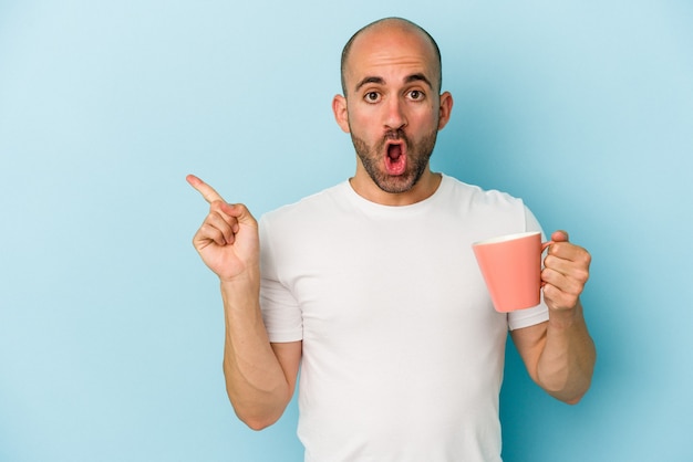 Photo young bald man holding a mug isolated on blue background  pointing to the side