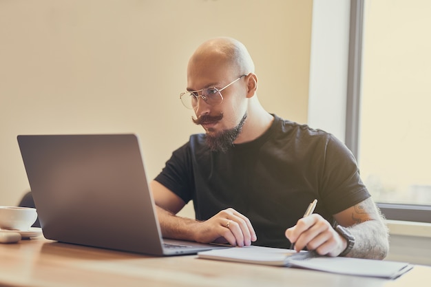 Young bald man working on laptop sitting at desk studying doing something notes distance learning