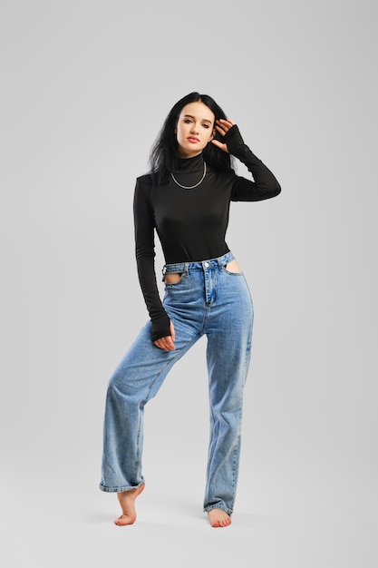 Young barefoot woman posing in studio in black bodysuit with long sleeves and boyfriend jean over grey background