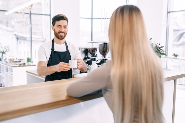 A young barista in a black apron smiles as he brings a cup of coffee to the girl behind the counter Service in a cafe